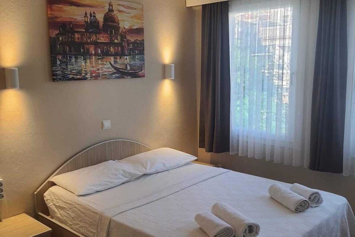 A hotel room with a bed, a nightstand, a painting of a cityscape, and a window with curtains.