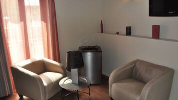 A living room with two beige armchairs, a small round table with a lamp, a compact refrigerator, and a flat-screen TV mounted on the wall.
