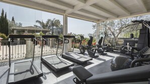 Un gimnasio al aire libre techado con equipamiento de ejercicio, incluyendo cintas de correr y máquinas de pesas, y vistas a un jardín y edificios al fondo.
