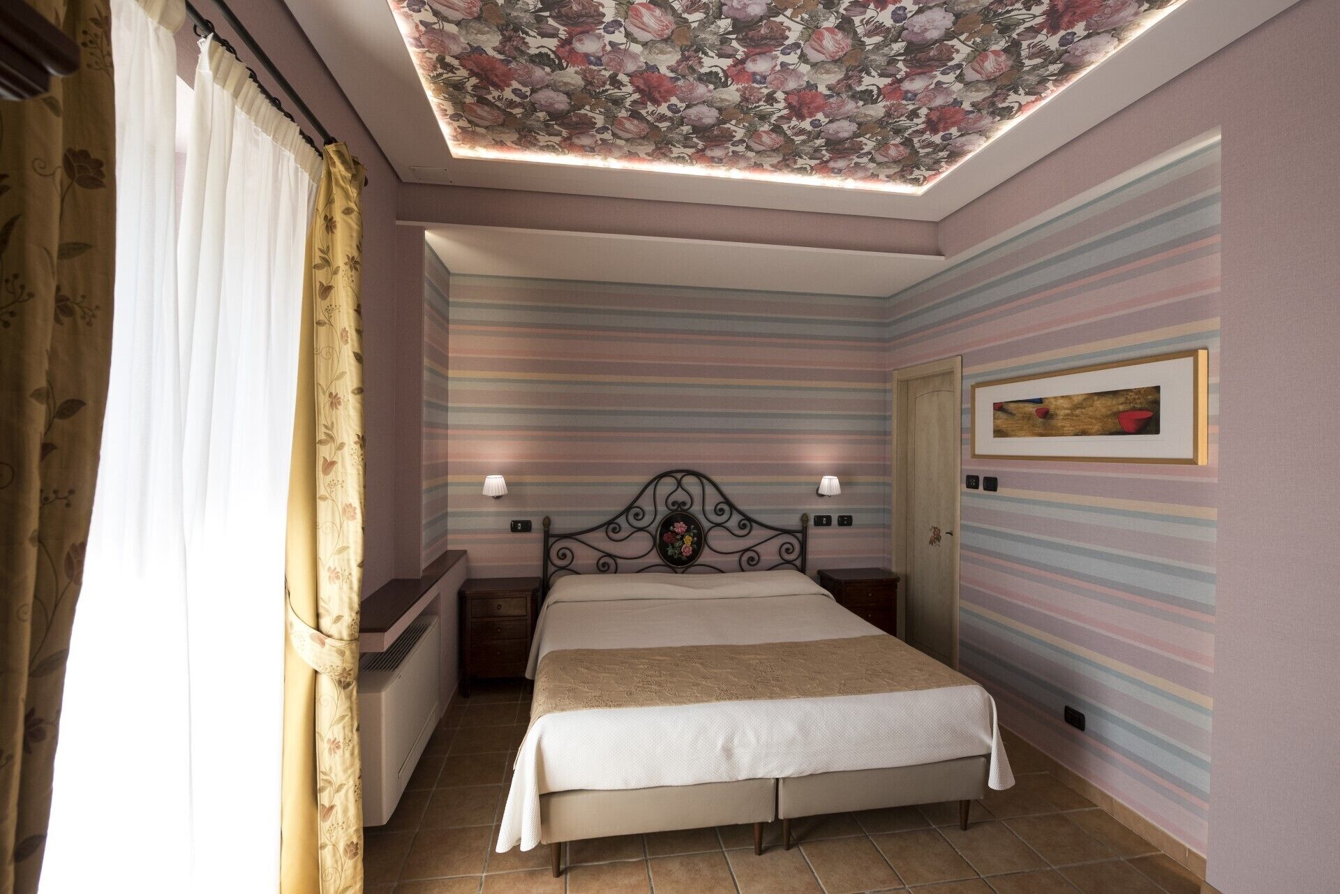 A bedroom with a bed, two bedside lamps, a floral ceiling, striped walls, and a door.