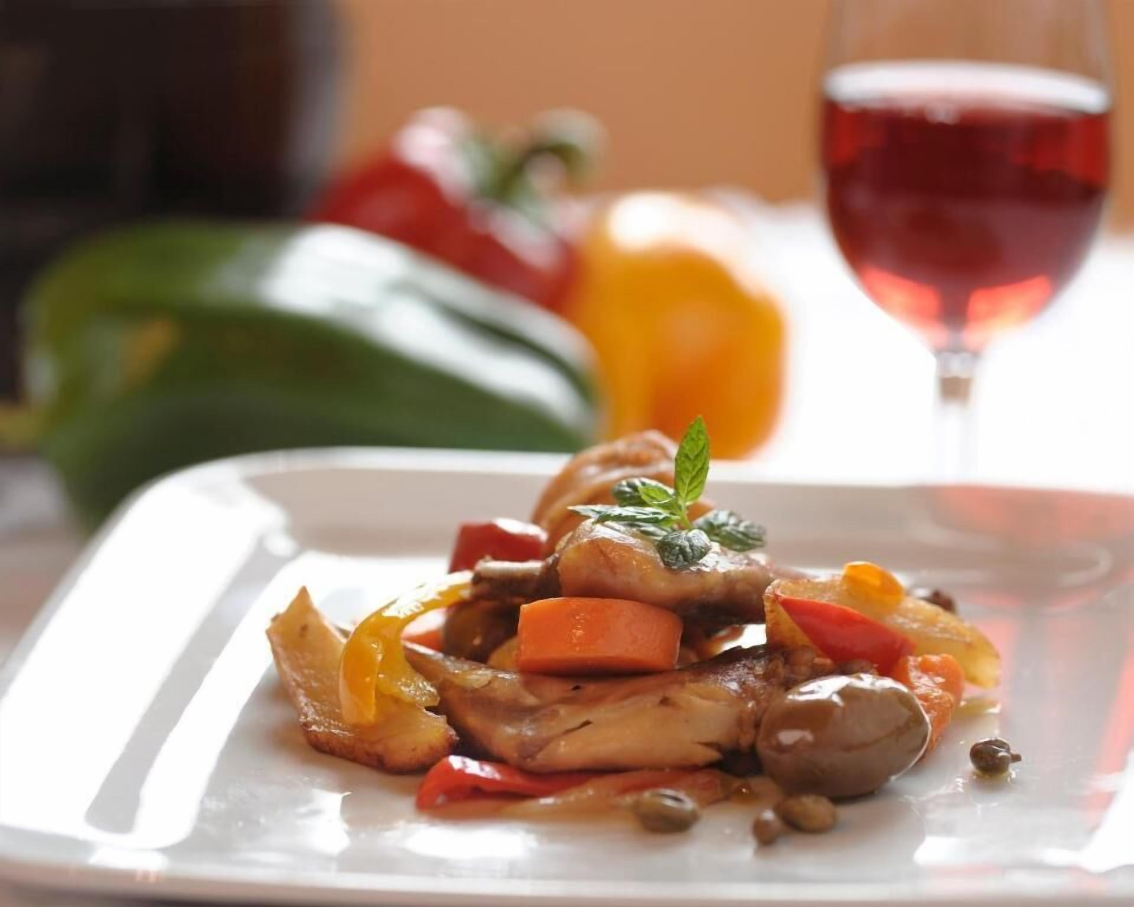 Un plato cuadrado con un guiso de verduras y carne, decorado con una ramita de hierba, acompañado de una copa de vino tinto y un fondo desenfocado de vegetales.