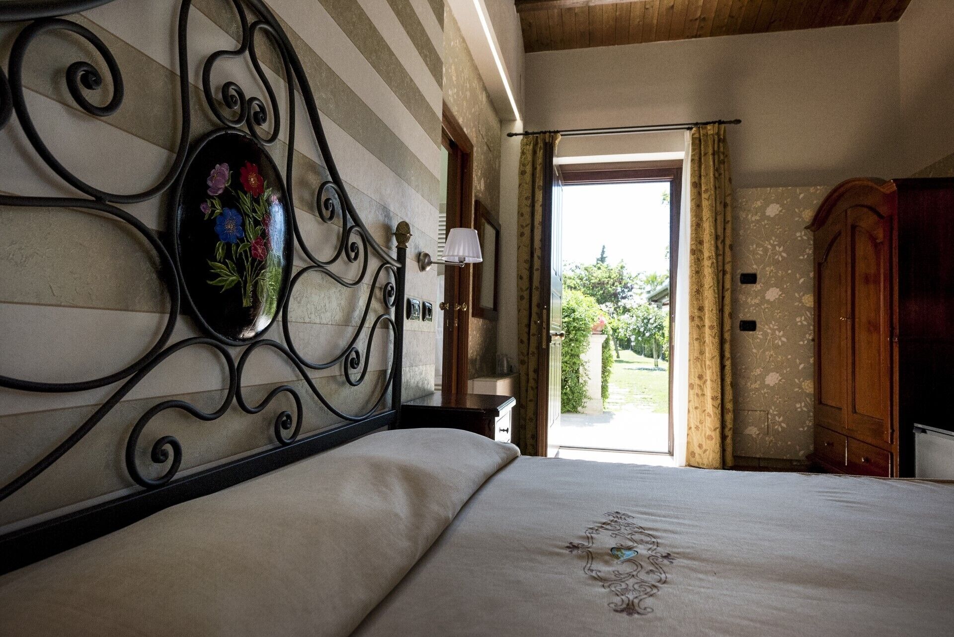 A bedroom with a bed, a decorative mirror, a wooden wardrobe, and a view of the outdoors.