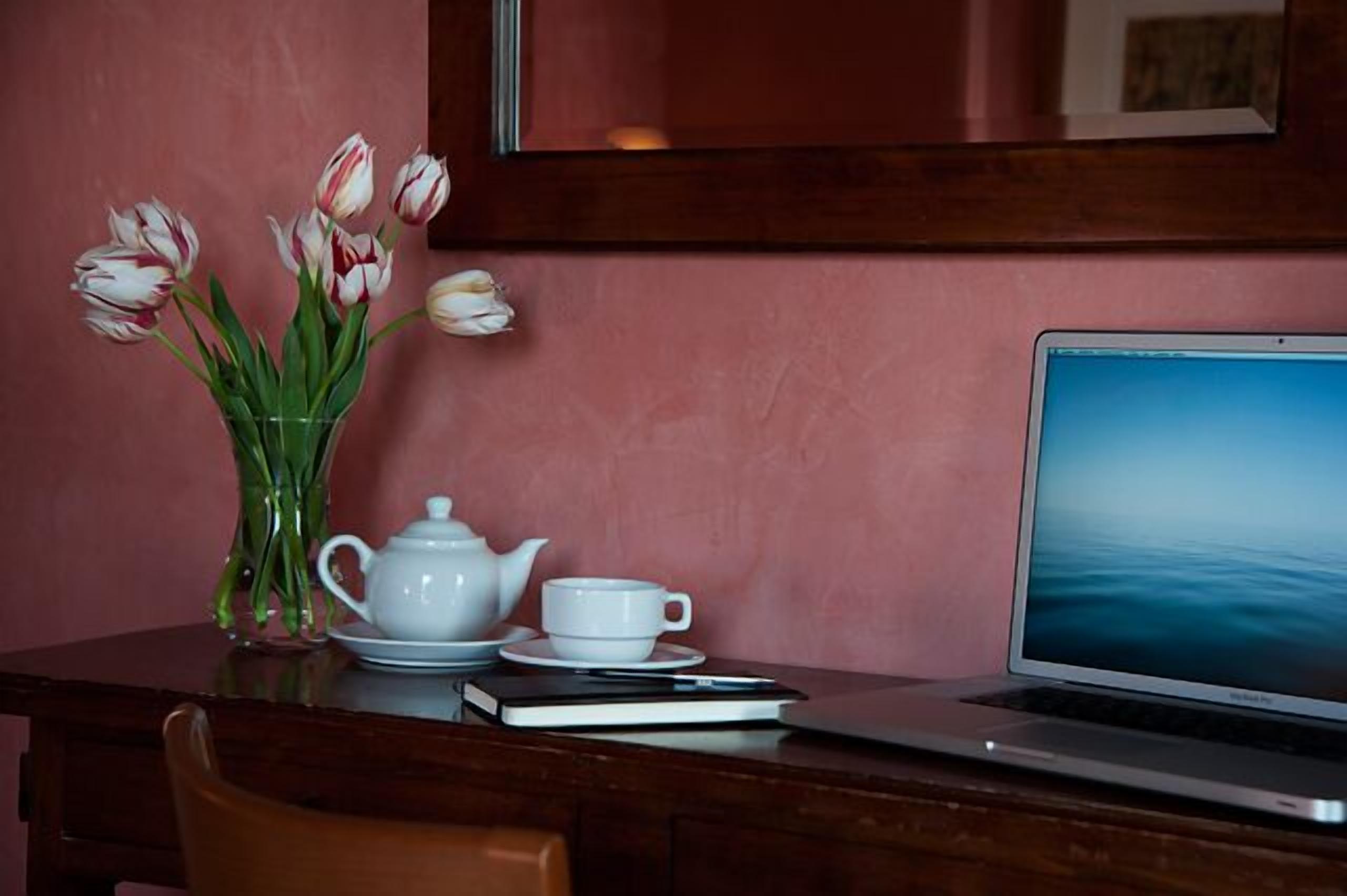 A laptop on a wooden table with a vase of tulips, a teapot, and a cup on the table.