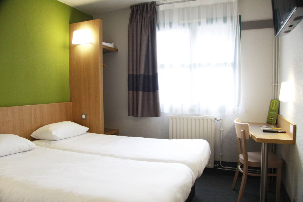 A hotel room with two beds, a desk, a chair, a radiator, and a window with curtains.