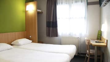 Habitación de hotel con dos camas, un escritorio, una silla, un radiador y una ventana con cortinas.