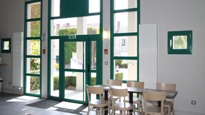 A room with a table and chairs, a door with a green frame, and a window with a view of buildings outside.