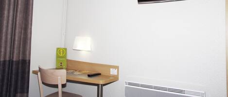 A hotel room with a flat-screen TV mounted on the wall, a desk with a chair, a lamp, and a heater.