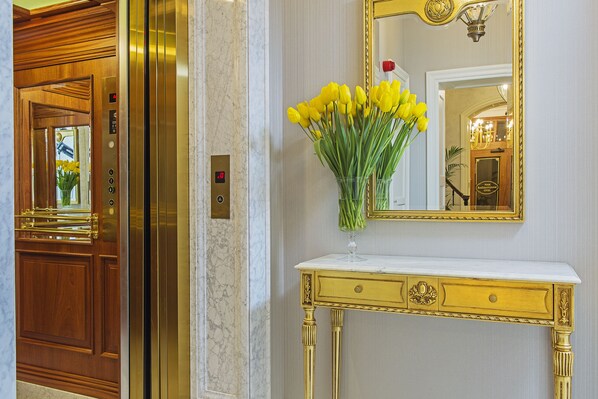 Porta de elevador em mármore, espelho com moldura dourada e mesa consola branca com um vaso de flores amarelas.