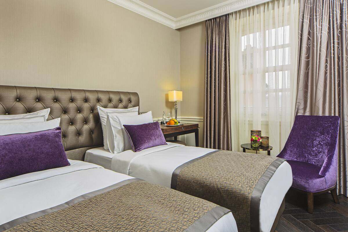 A hotel room with two beds, a nightstand with a lamp, a chair, and a view of the outdoors through curtains.