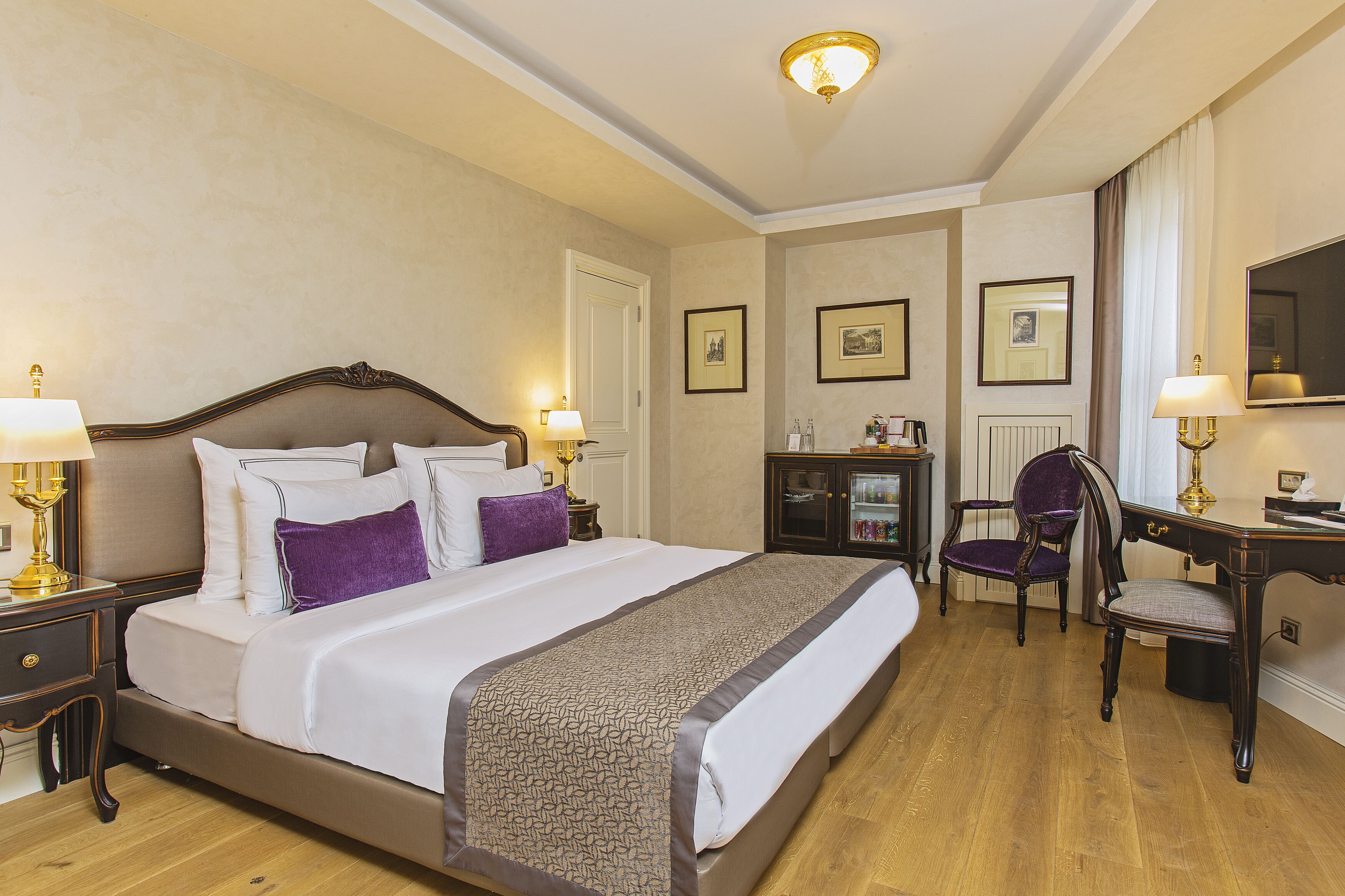 A hotel room with a large bed, two bedside tables with lamps, a desk with a chair, and framed pictures on the walls.