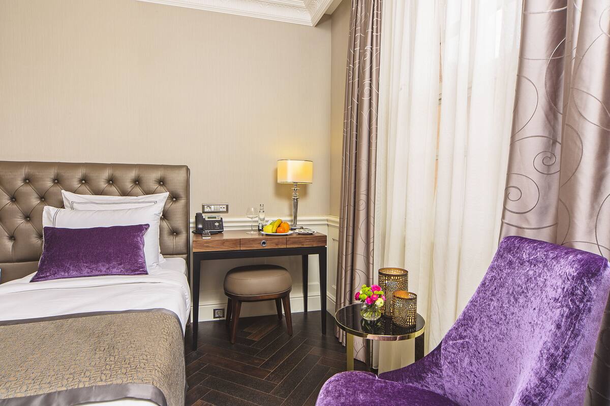 A hotel room with a large bed, a desk with a lamp, a chair, and a small table with a vase of flowers.