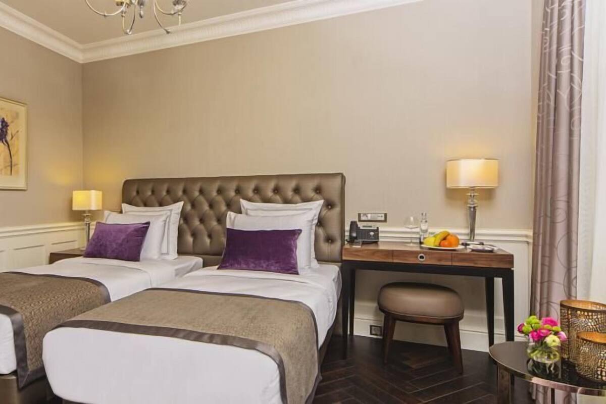 A hotel room with two single beds, a nightstand with a lamp, a small table with a fruit bowl, and a chair.
