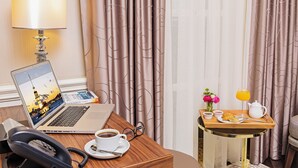 Habitación de hotel con una laptop sobre un escritorio, un teléfono, una taza de café y una bandeja con alimentos para el desayuno, además de cortinas.