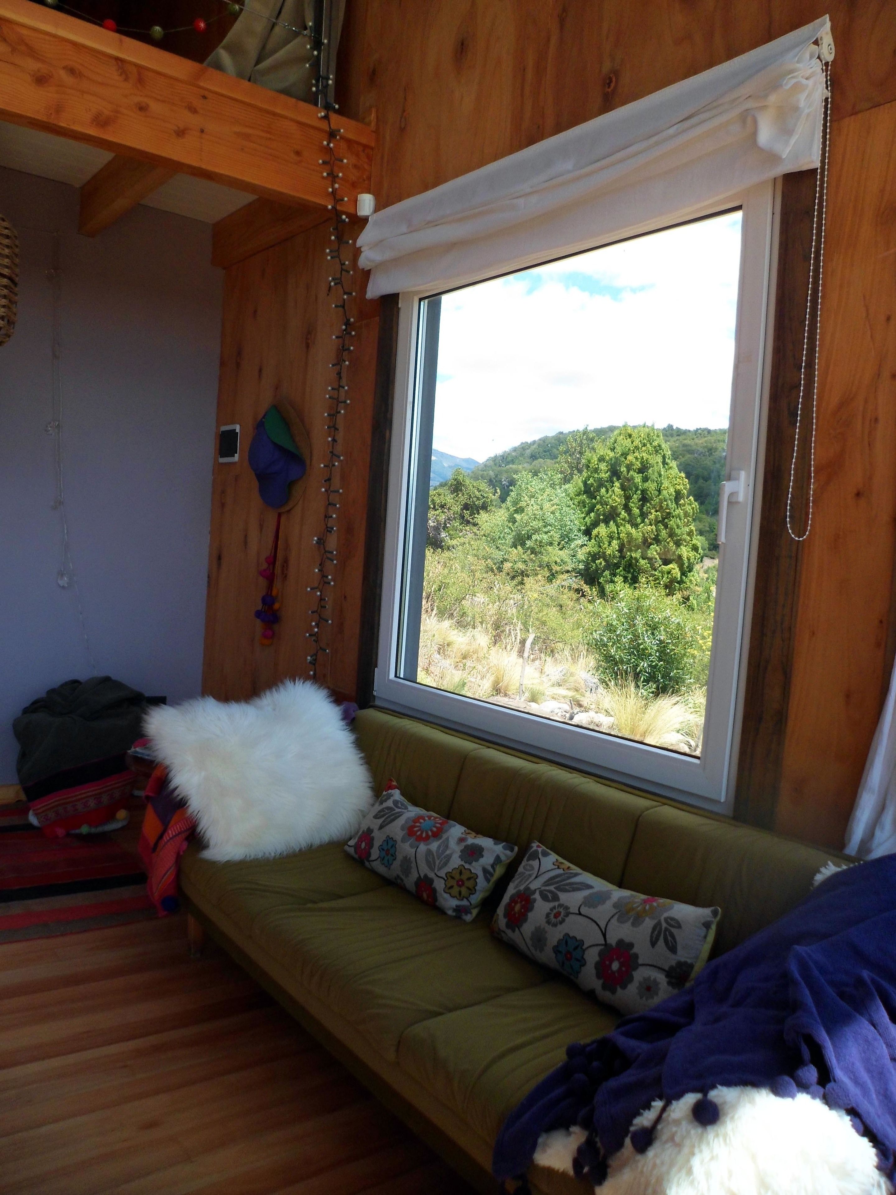 Una habitación acogedora con detalles de madera, un sofá verde con cojines estampados, un ventanal con vistas a colinas verdes y un adorno colgante.
