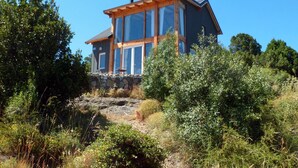 A two-story house with a stone foundation, large windows, and a wooden roof. - Un Vistón Modern Mountain Home (San Martín de los Andes)
