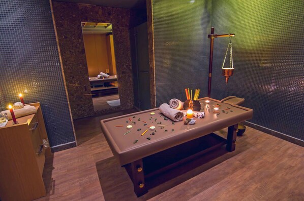 Mesa de masajes con velas, una toalla y diversas herramientas de masaje en un spa con espejo y pared texturizada.