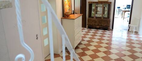 A room with a checkered floor, a white staircase, a wooden cabinet with a mirror, and a door leading to another room.
