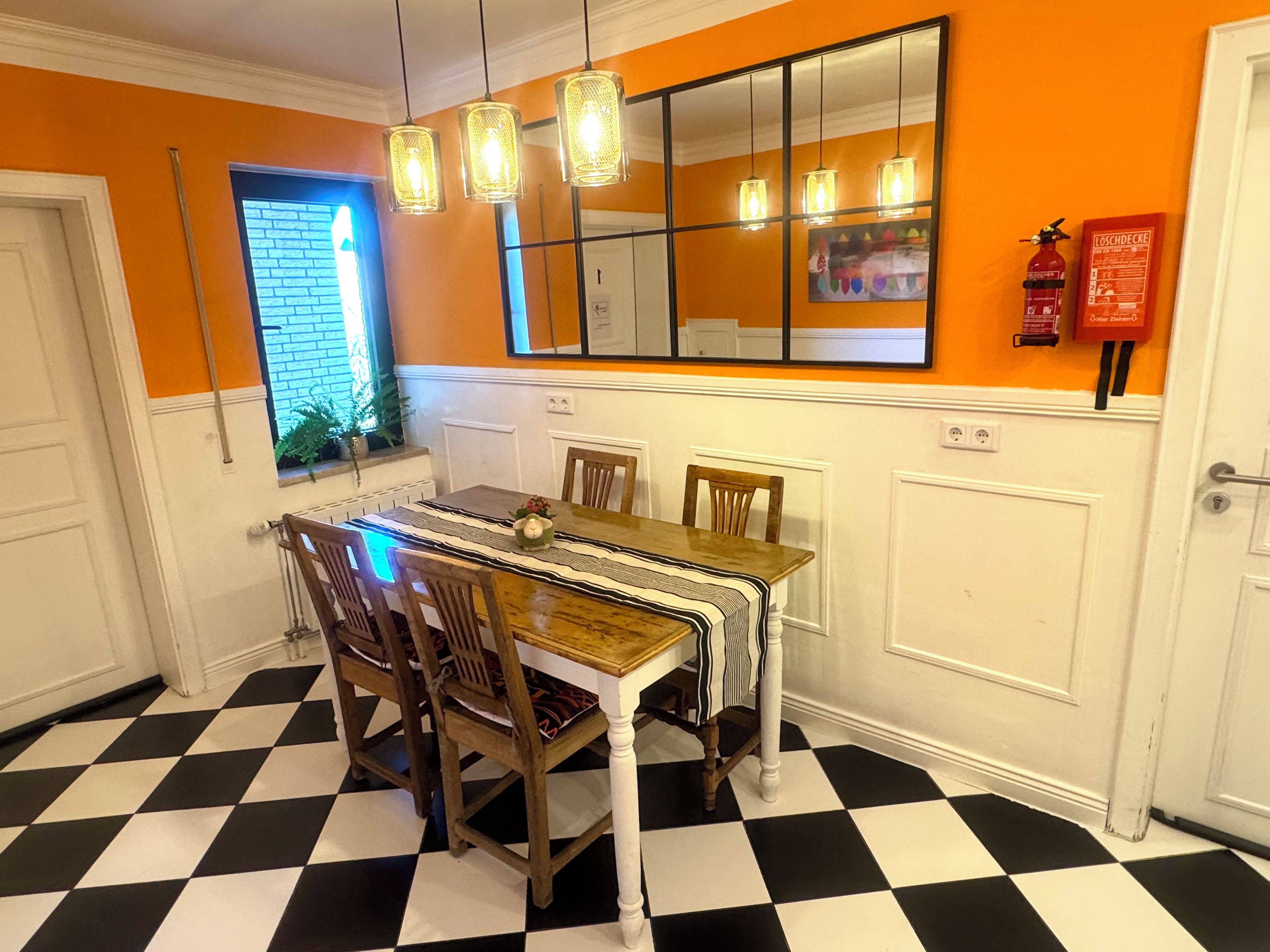 A dining area with a checkered floor, wooden chairs, a table with a striped tablecloth, pendant lights, a mirror, and a fire extinguisher.