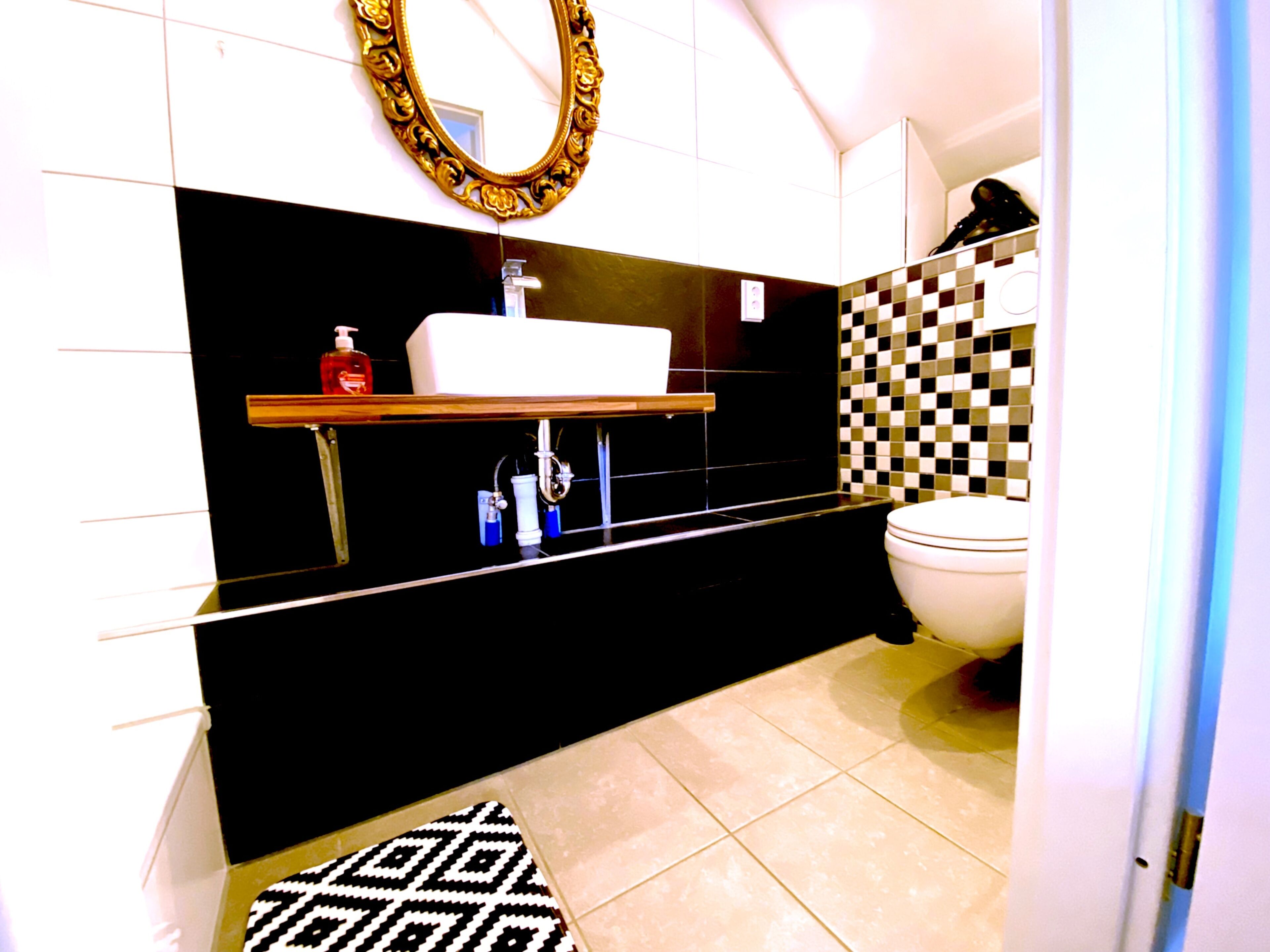 Ein modernes Badezimmer mit schwarz-weiß kariertem Fliesenspiegel, einem weißen Waschbecken und einem goldfarbenen Spiegel darüber sowie einer weißen Toilette.