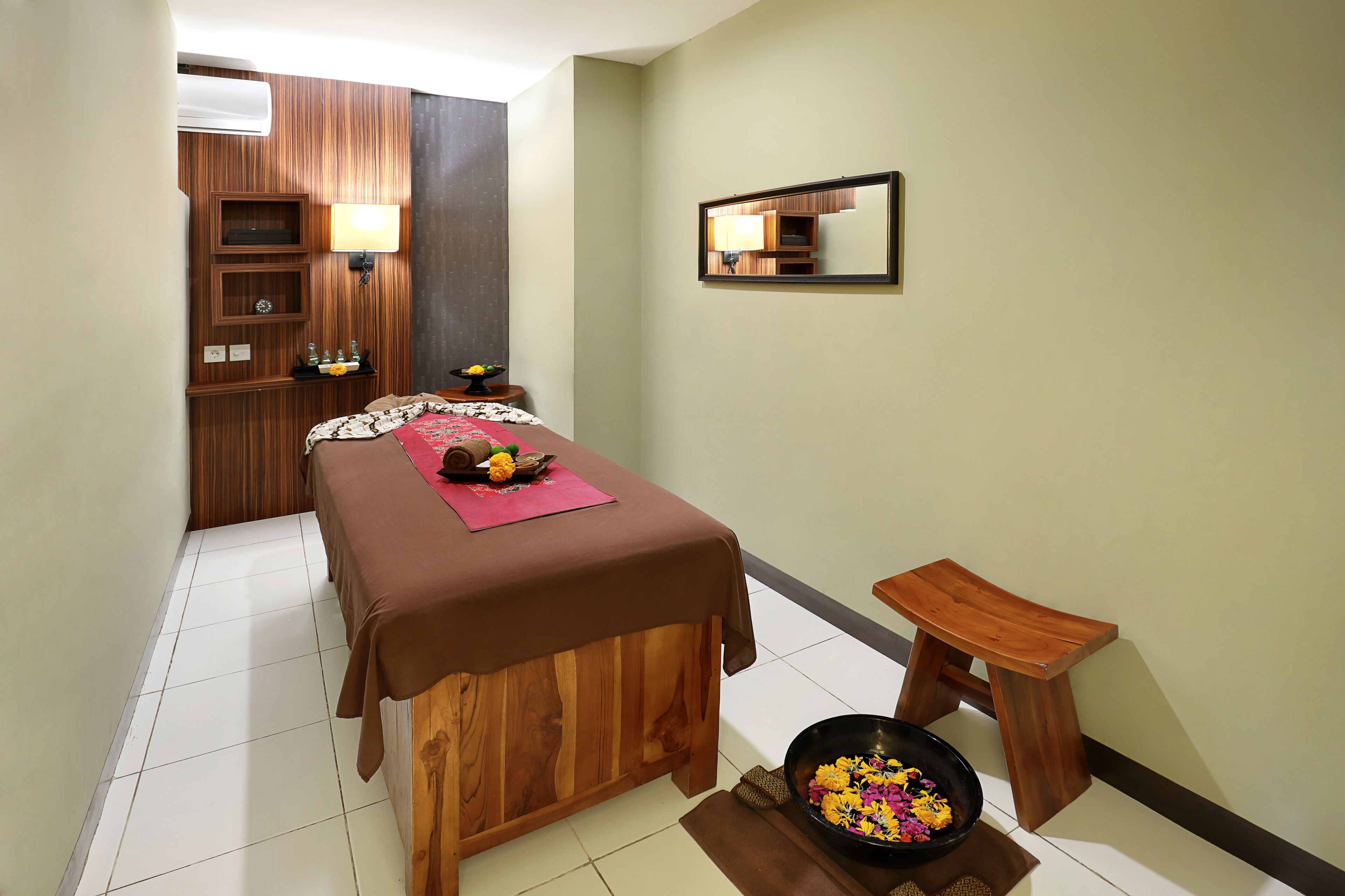 A massage room with a wooden massage table, a small stool, a bowl of flowers, and a mirror on the wall.