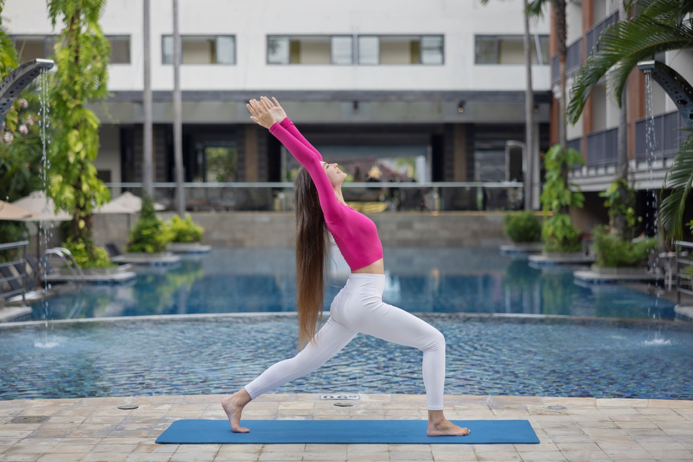 A person performing a yoga pose on a mat by a pool with a building in the background.