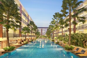 A pool area with a long, narrow pool, surrounded by palm trees and lounge chairs, with a multi-story building in the background.