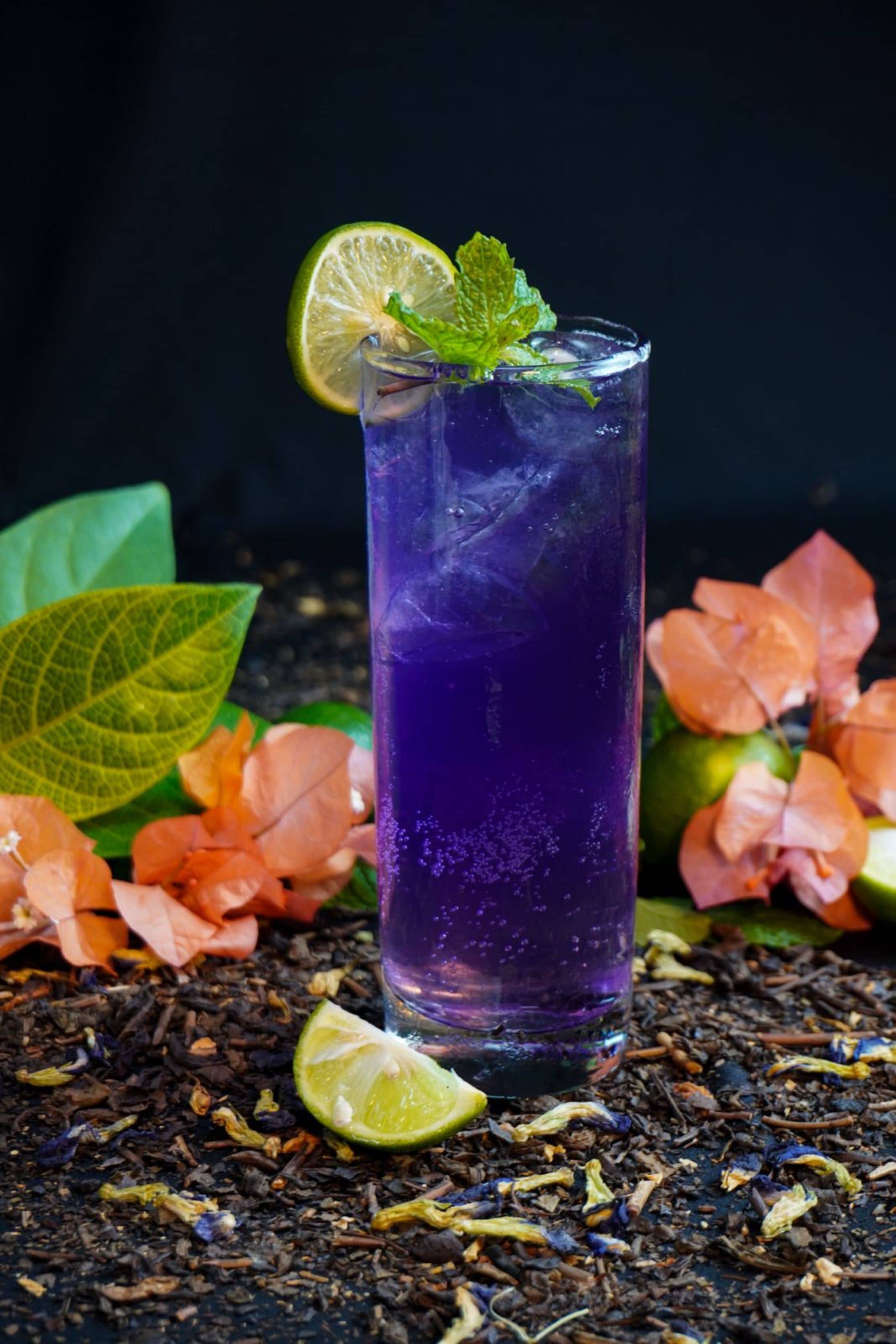 A tall glass of purple beverage with a lime slice and mint garnish, surrounded by tea leaves and flowers on a dark surface.