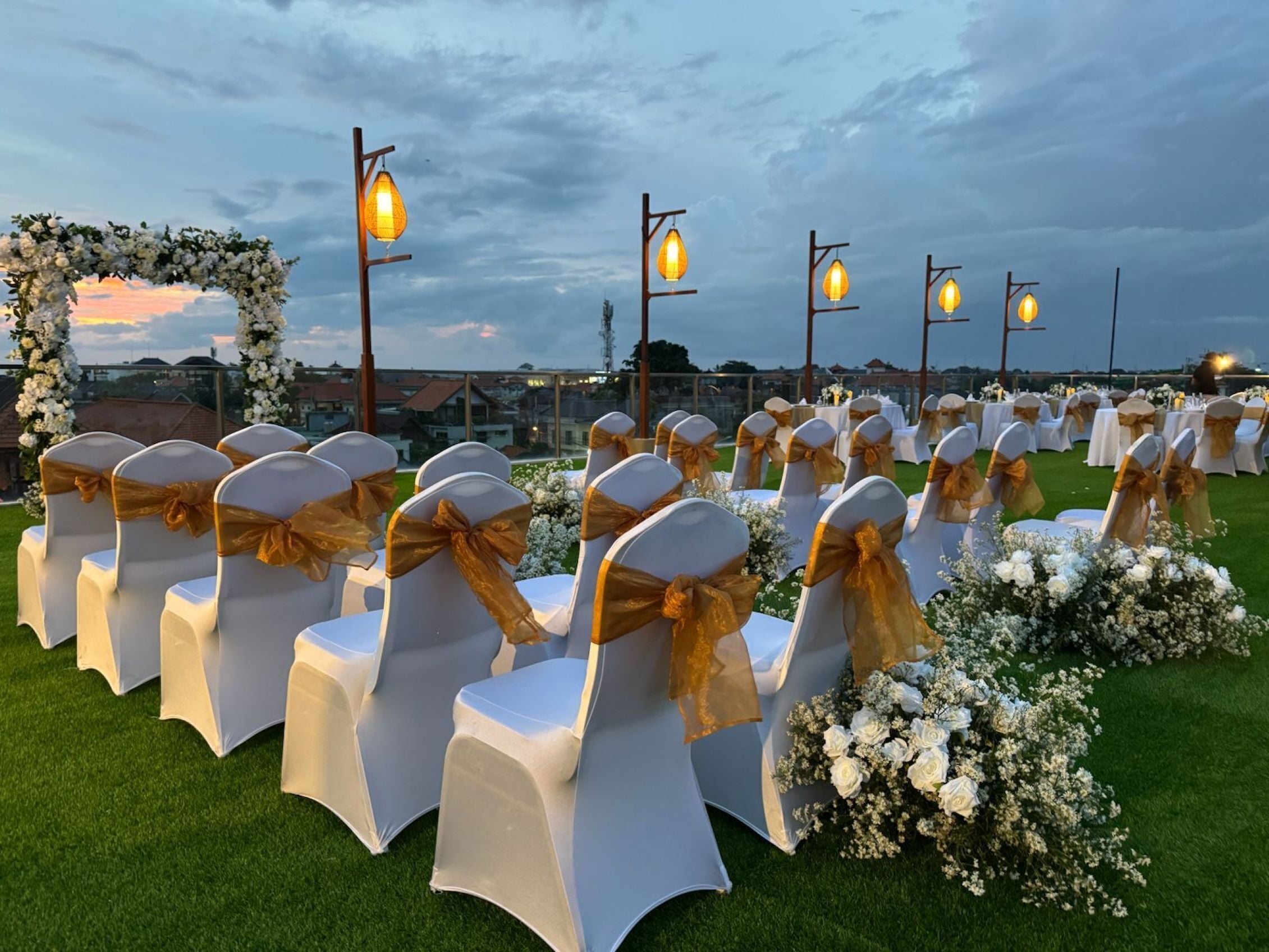 Rows of white chairs with gold sashes, arranged in a semi-circle with a floral arch and hanging lanterns.