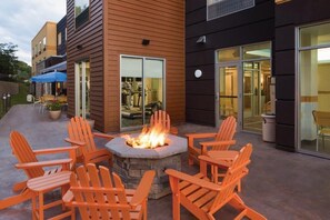 An outdoor seating area with a fire pit and Adirondack chairs, adjacent to a building with large windows and a door.