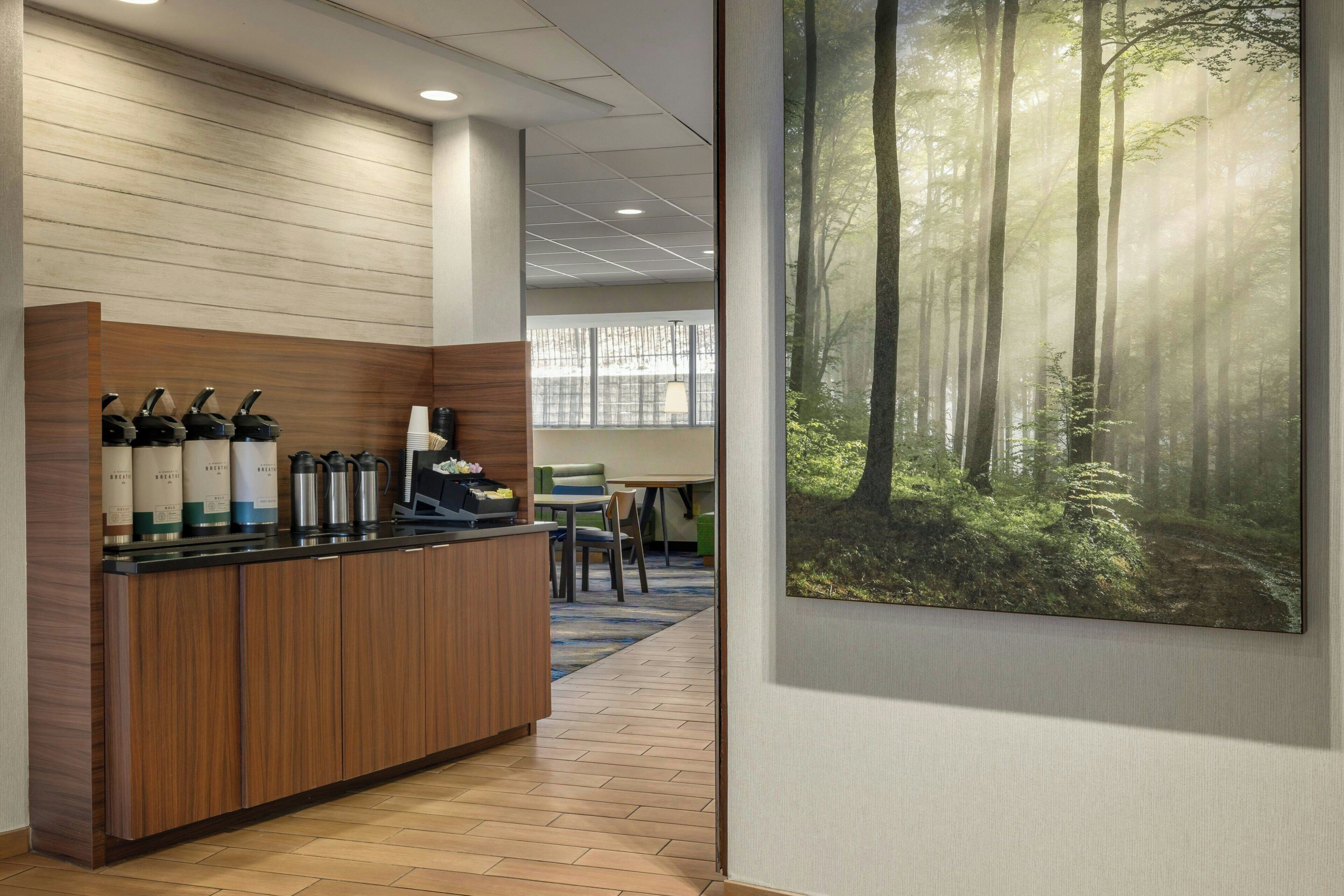 A reception area with a wooden counter, coffee makers, and a large forest-themed painting on the wall.