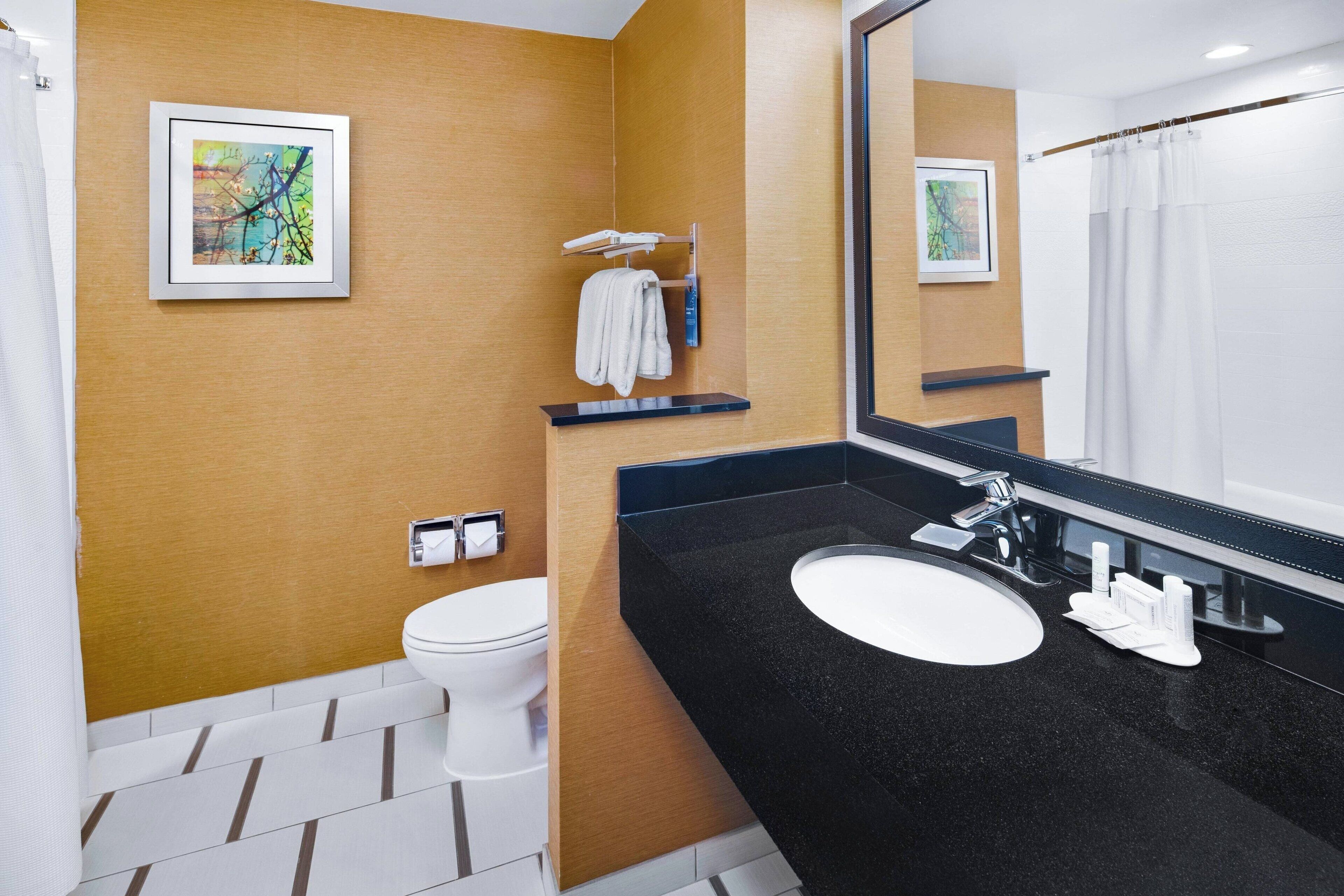 A modern bathroom with a black countertop, white sink, and a large mirror. There is a towel rack above the toilet and a framed abstract painting on the wall.