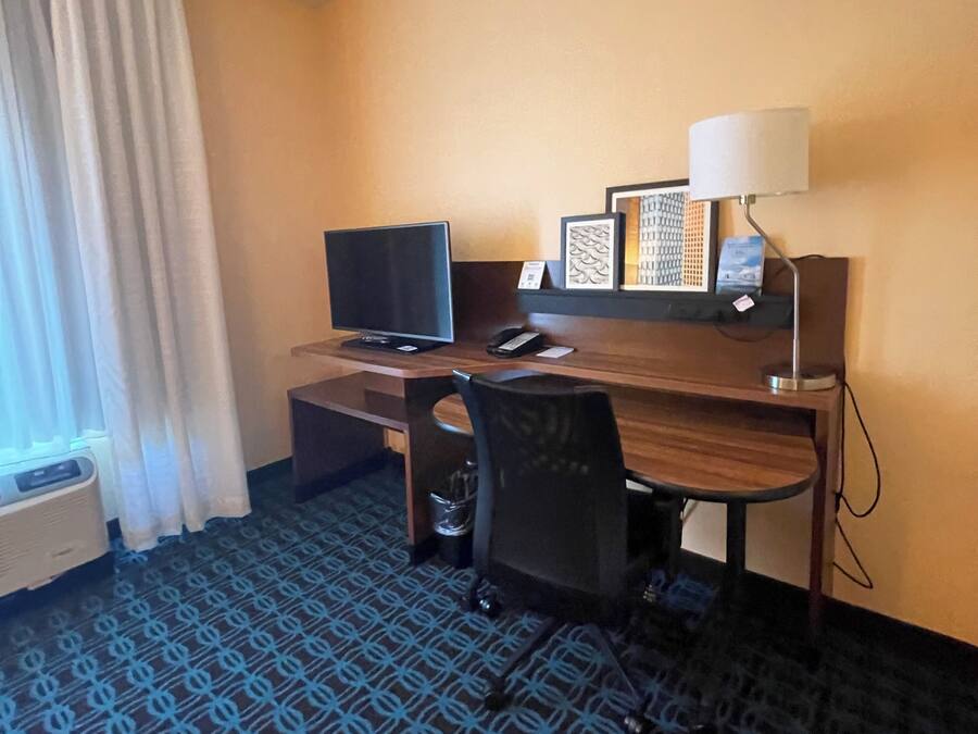 A hotel room with a desk, chair, television, and a patterned carpet.
