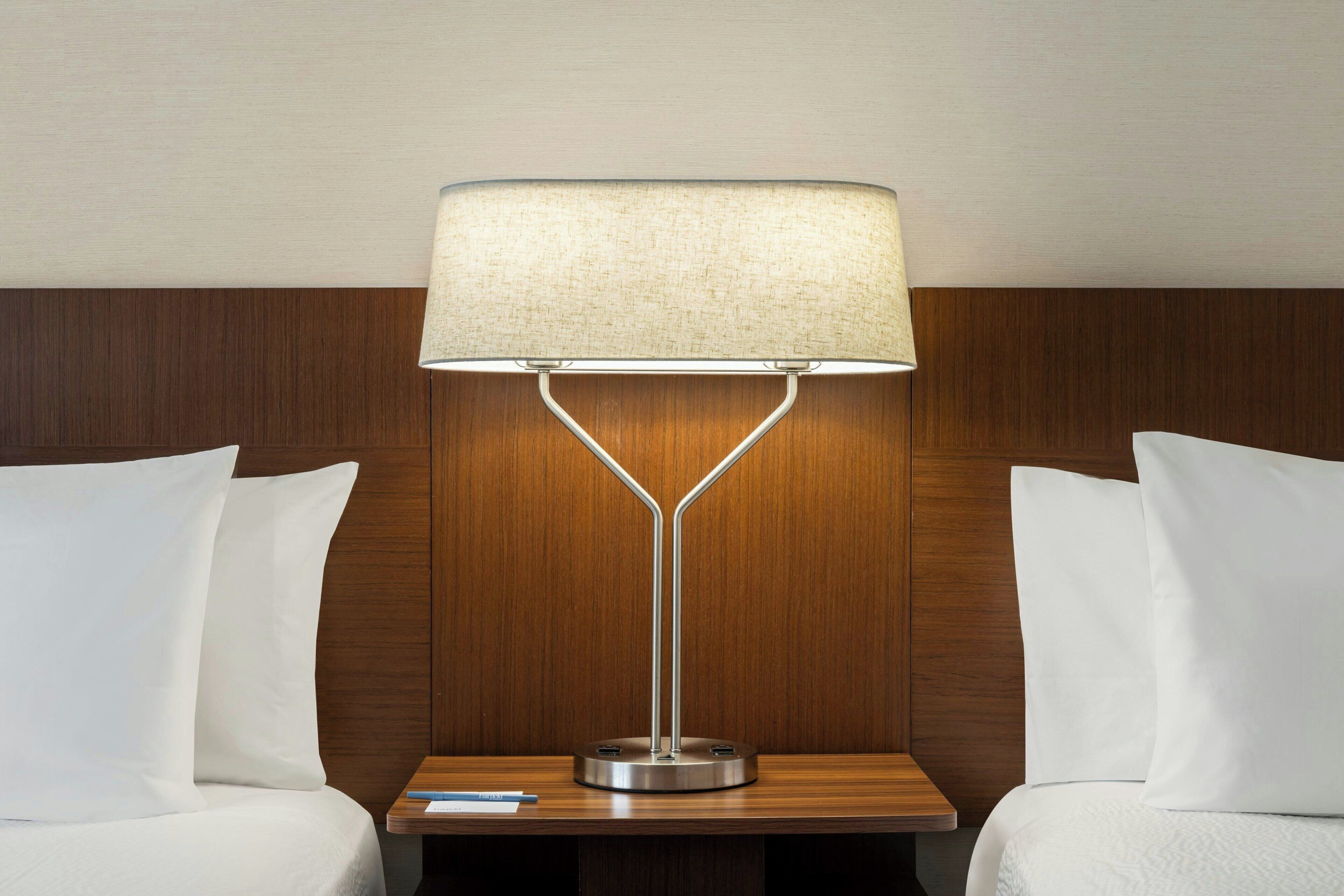 A hotel room with a wooden headboard, a bedside table with a lamp, and two white pillows.