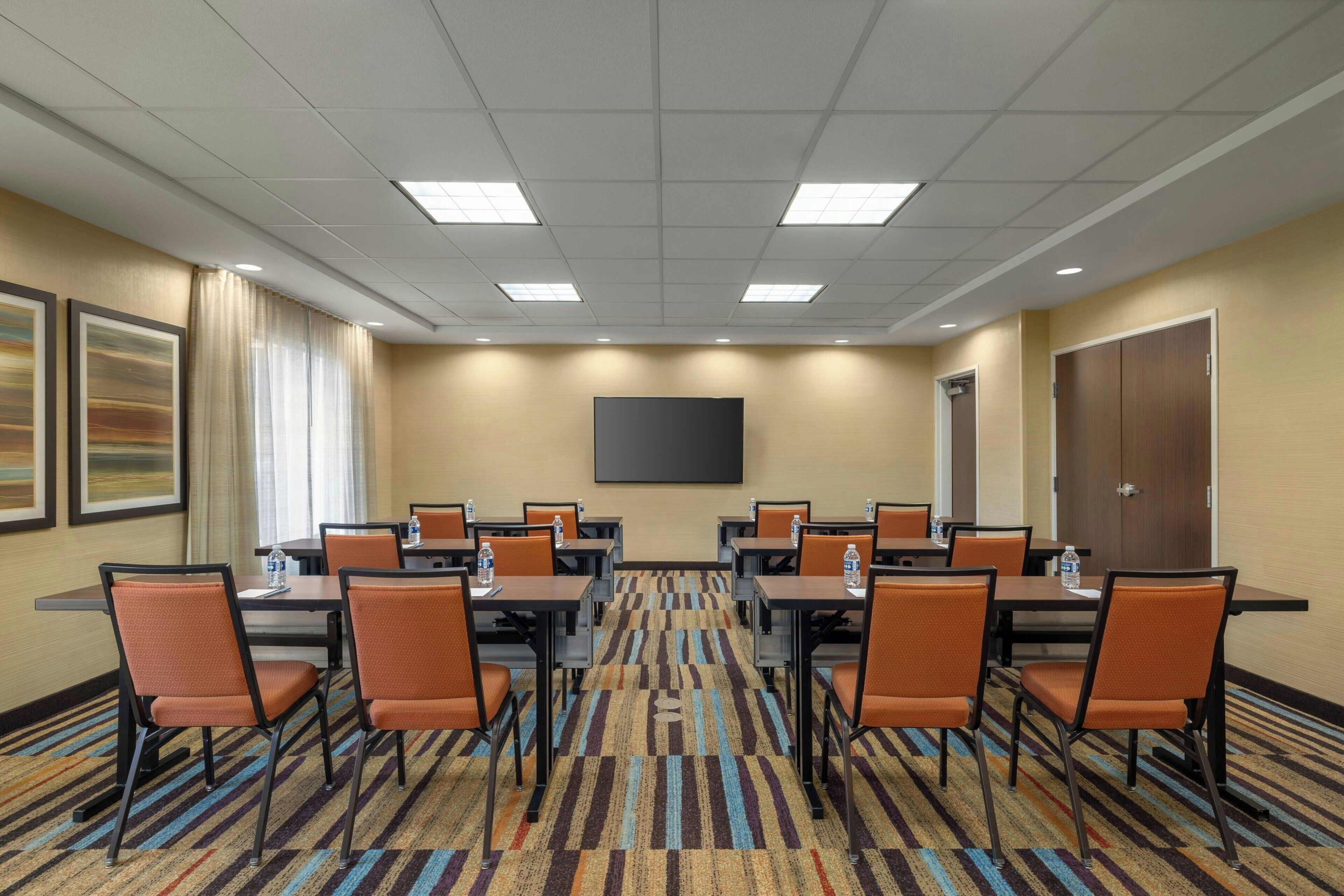 A conference room with a carpeted floor, tables, chairs, and a flat-screen TV on the wall.