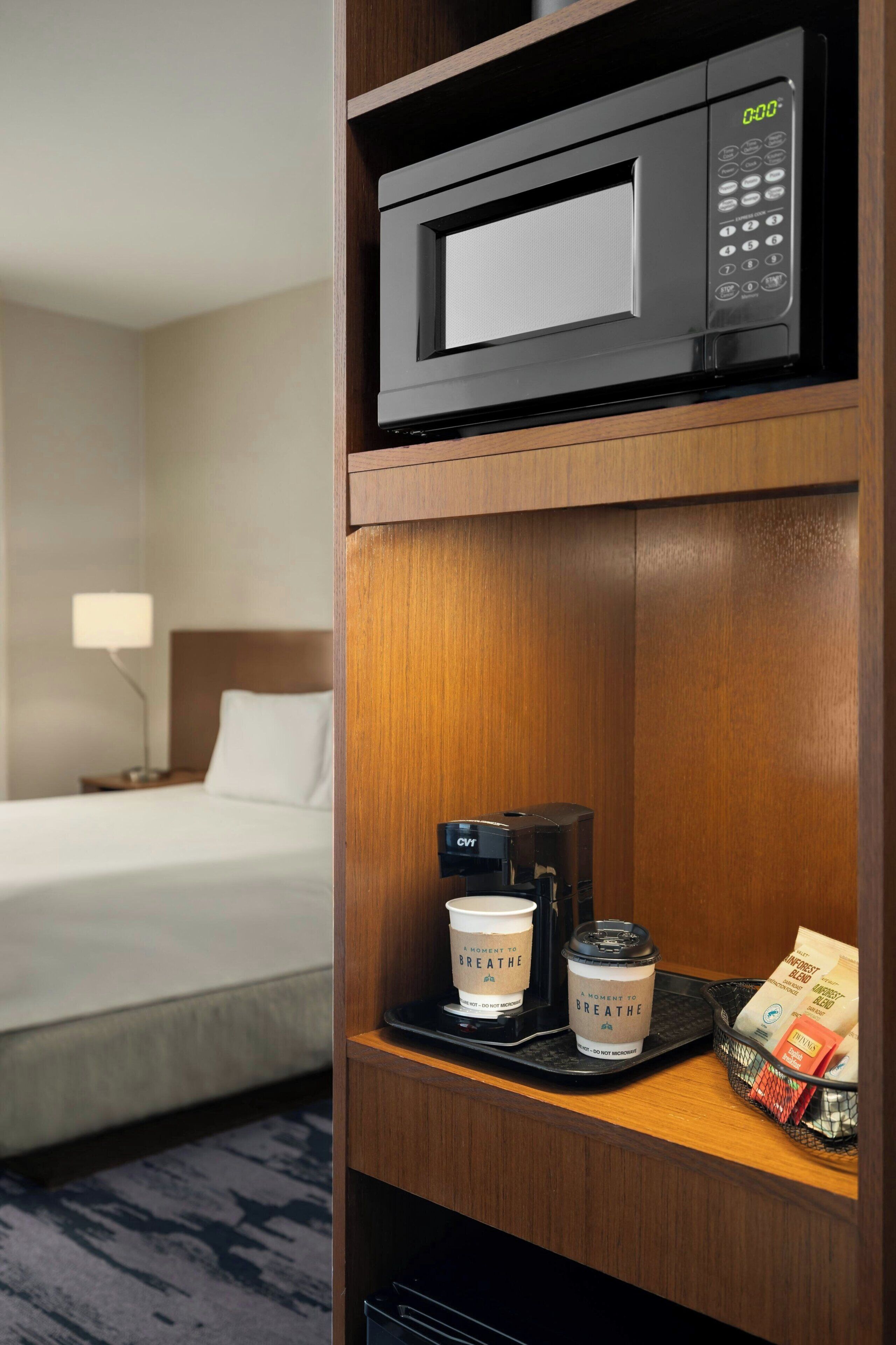 A hotel room with a microwave on a wooden shelf, a coffee maker, two coffee cups, and a basket of coffee pods.