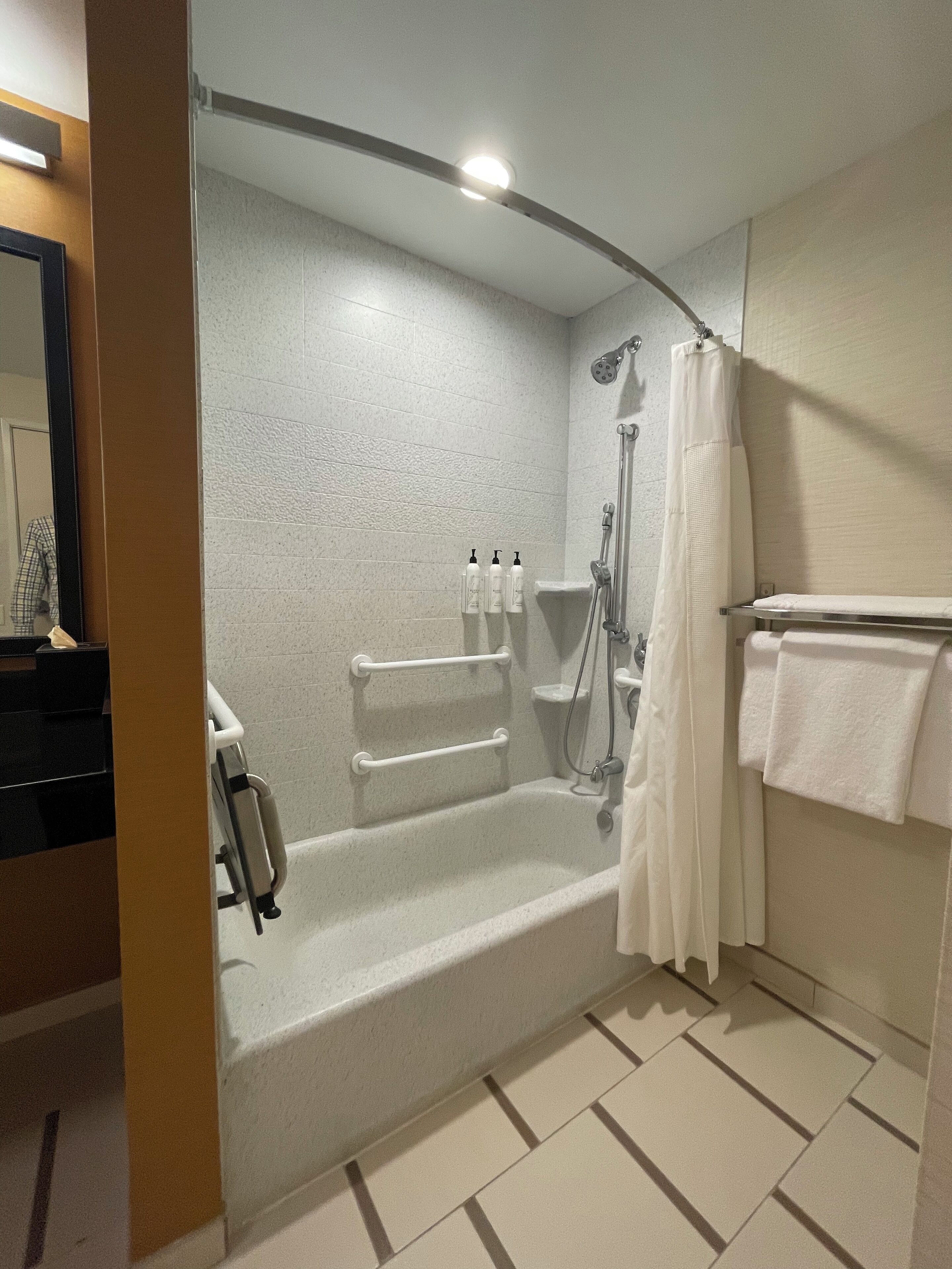 A modern bathroom with a bathtub, shower, and toilet. There are handrails and a towel rack.