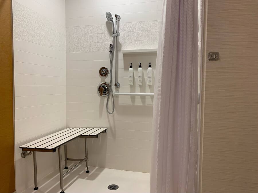 A small, white-tiled bathroom with a shower, a bench, and a shelf with toiletries.