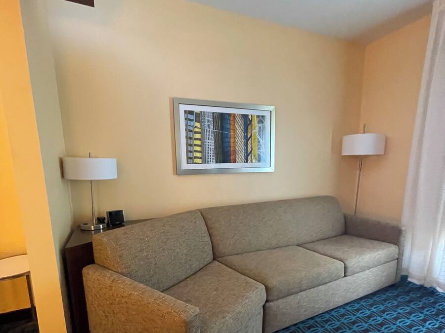A hotel room with a beige sofa, a framed picture on the wall, two floor lamps, and a patterned carpet.