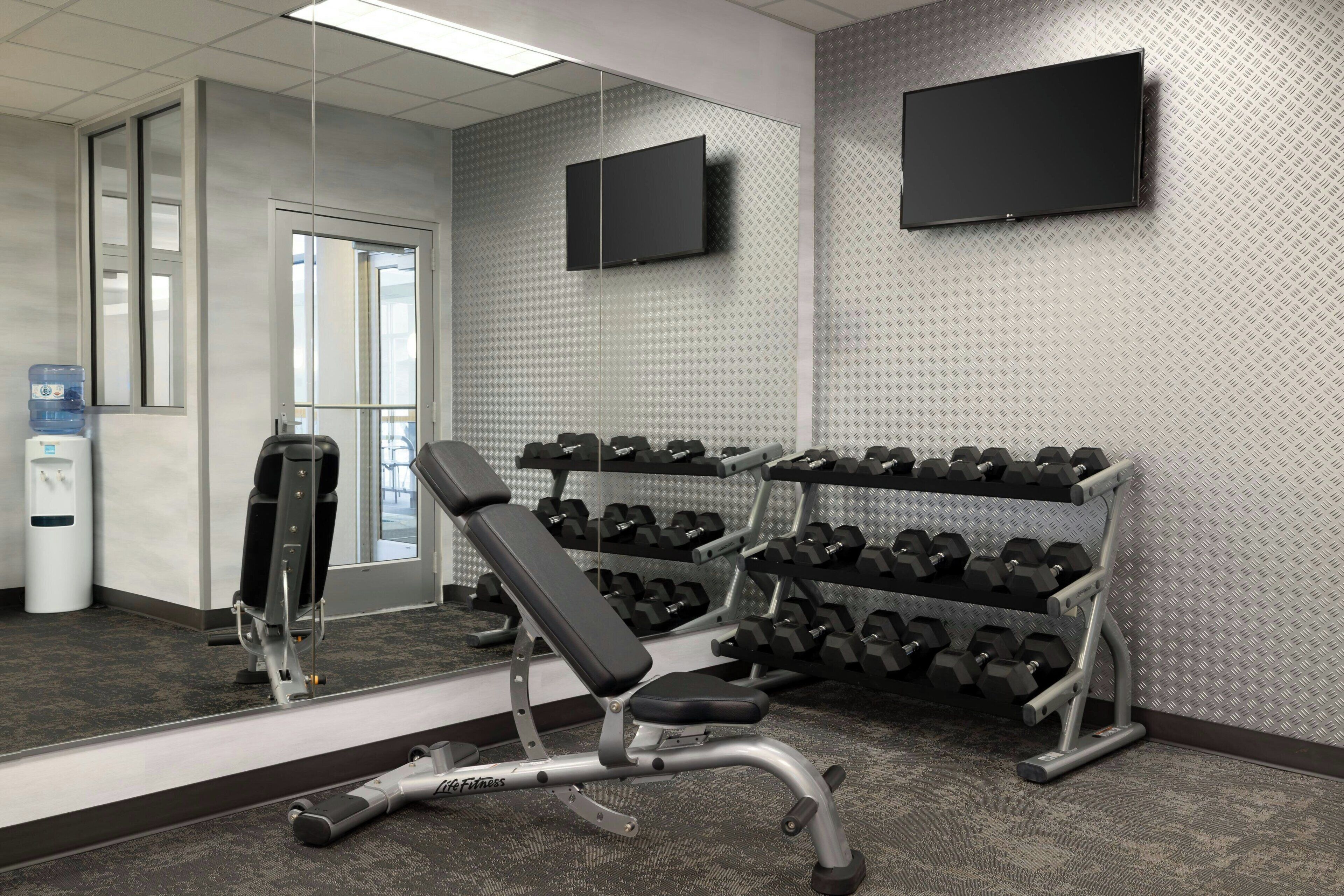 A gym with a bench, dumbbells, a mirror, and a TV on the wall.