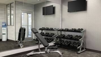 A gym with a bench, dumbbells, a mirror, and a TV on the wall.