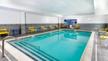 En indendørs swimmingpool med krystalklart vand, omgivet af gule og blå stole, og et flisegulv.