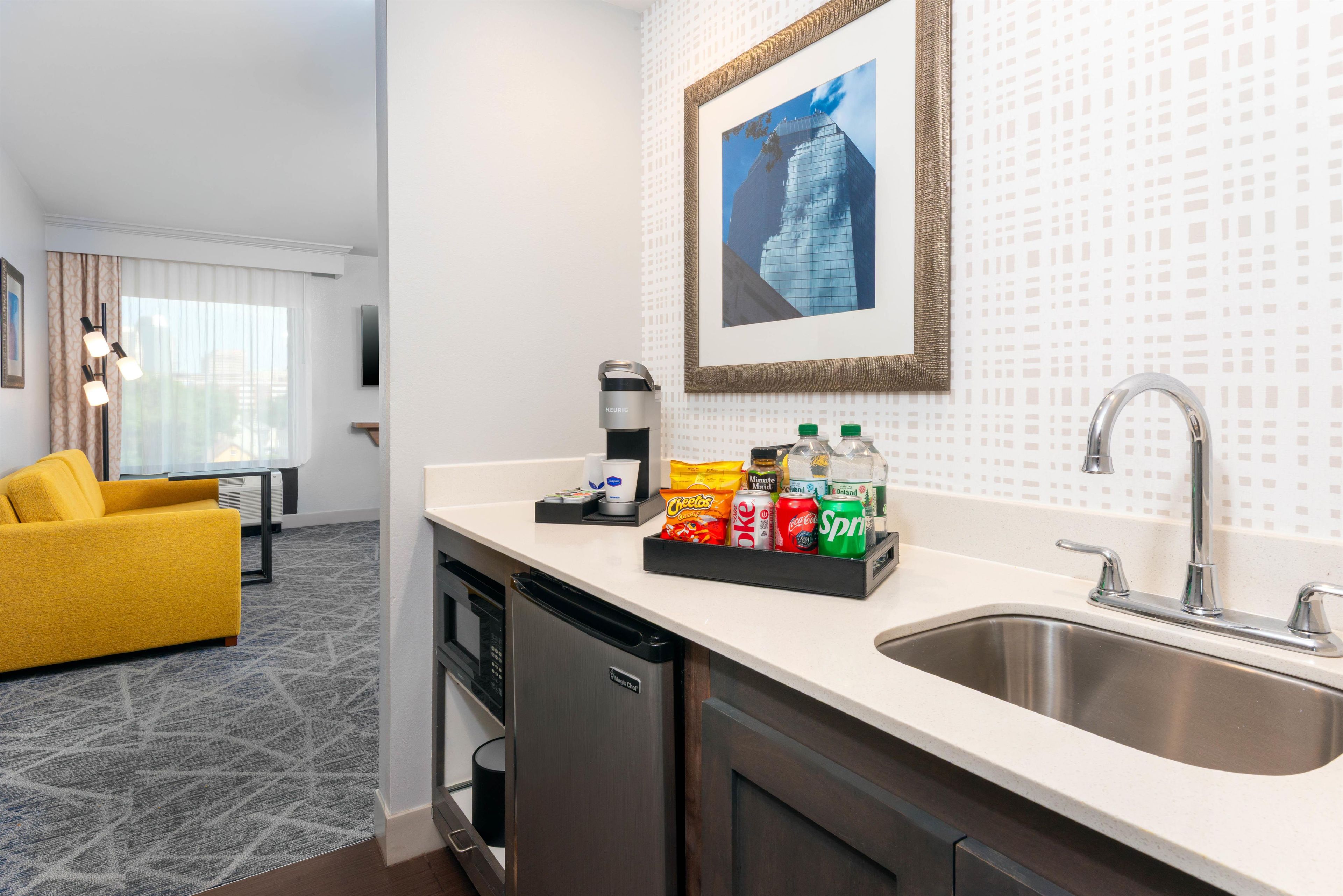 A hotel room with a kitchenette, a dining area with a yellow sofa, a large window with curtains, and a framed picture on the wall.