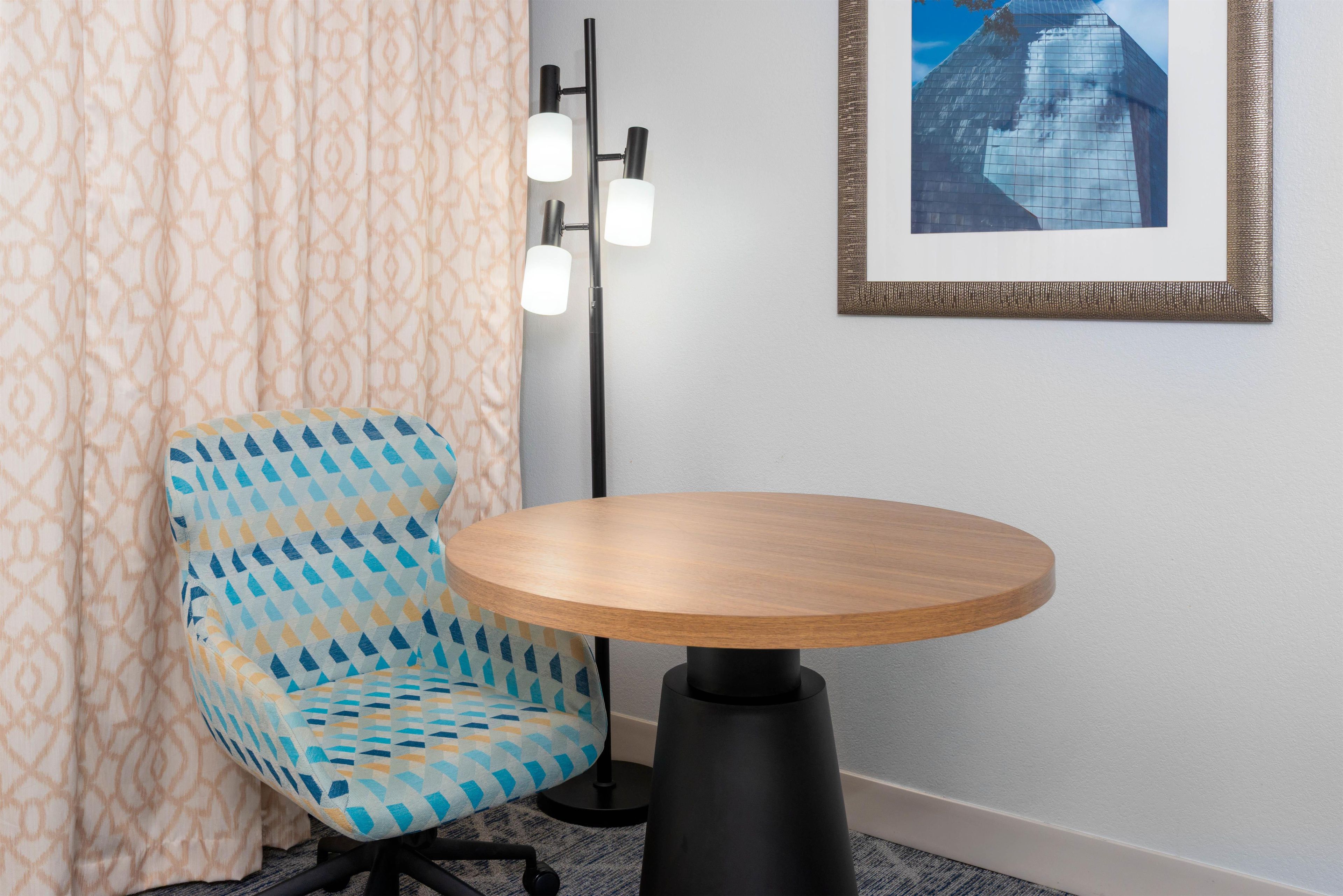 A room with a patterned chair, a round wooden table, a floor lamp, and a framed picture on the wall.