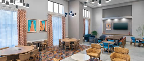 A hotel lobby with a large meeting table, several smaller tables, comfortable seating, artwork on the walls, and a flat-screen TV mounted on the wall.