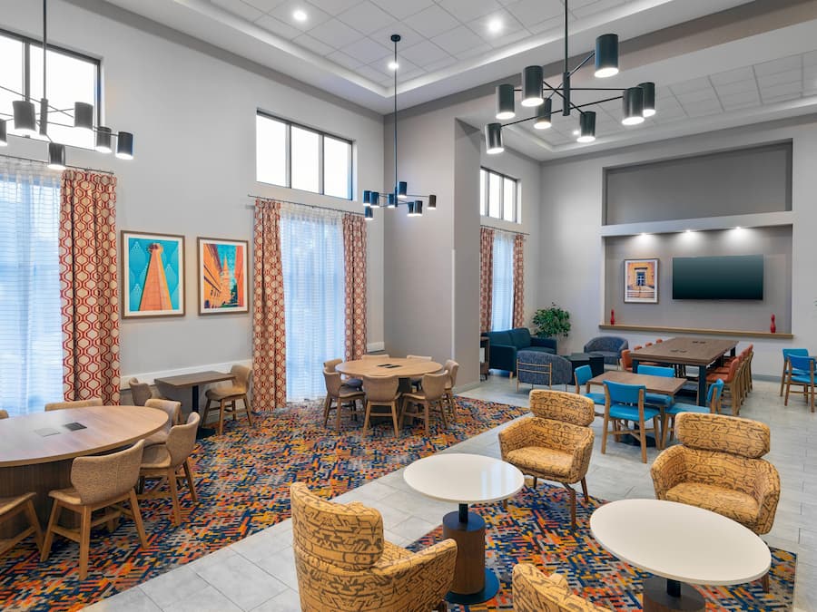 Un vestíbulo de hotel con una gran mesa de reuniones, varias mesas más pequeñas, asientos cómodos, obras de arte en las paredes y un televisor de pantalla plana montado en la pared.