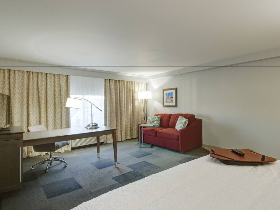 A hotel room with a desk, a red sofa, a bed, and a remote control on the bed.