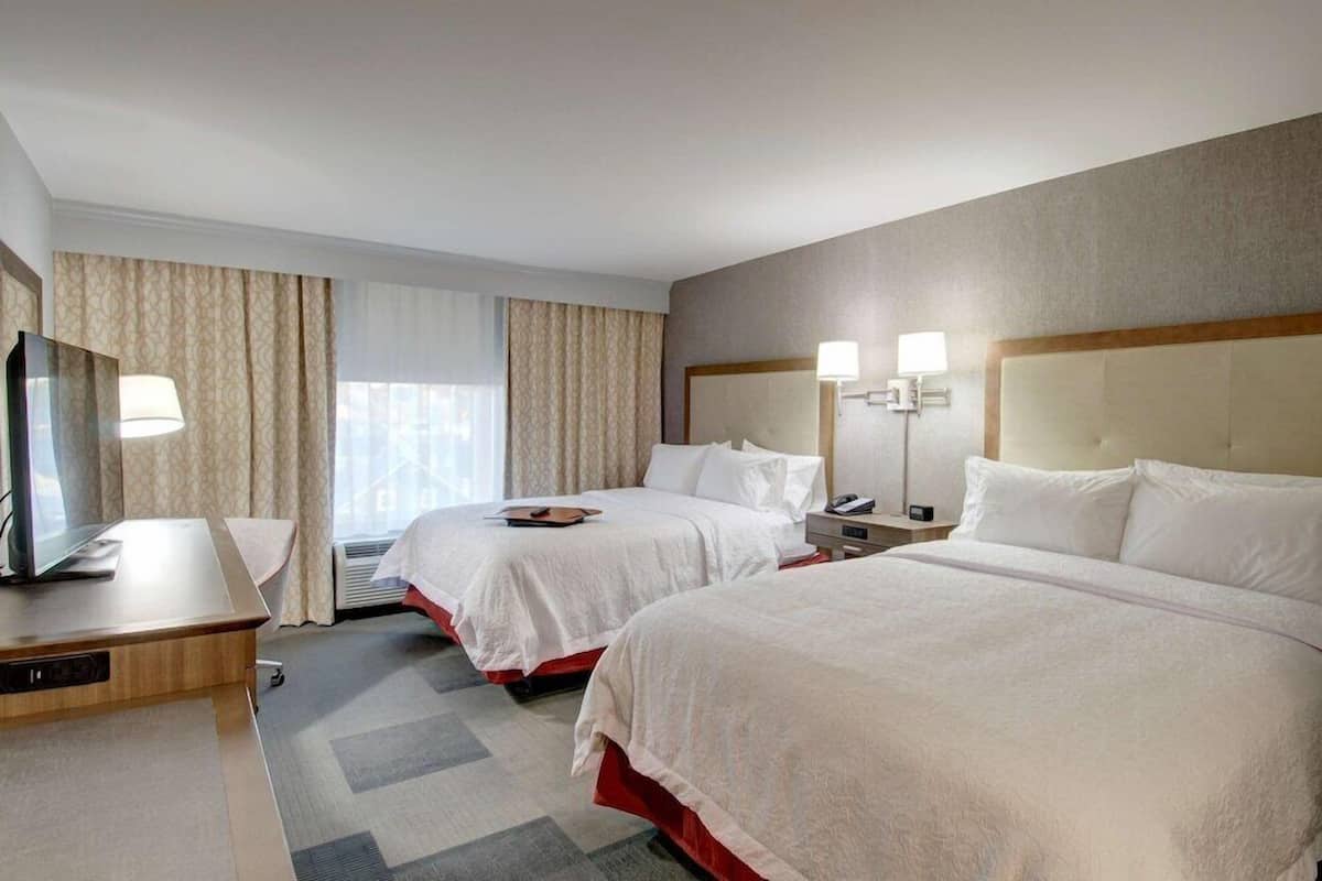 A hotel room with two beds, a desk, a TV, and a window with curtains.