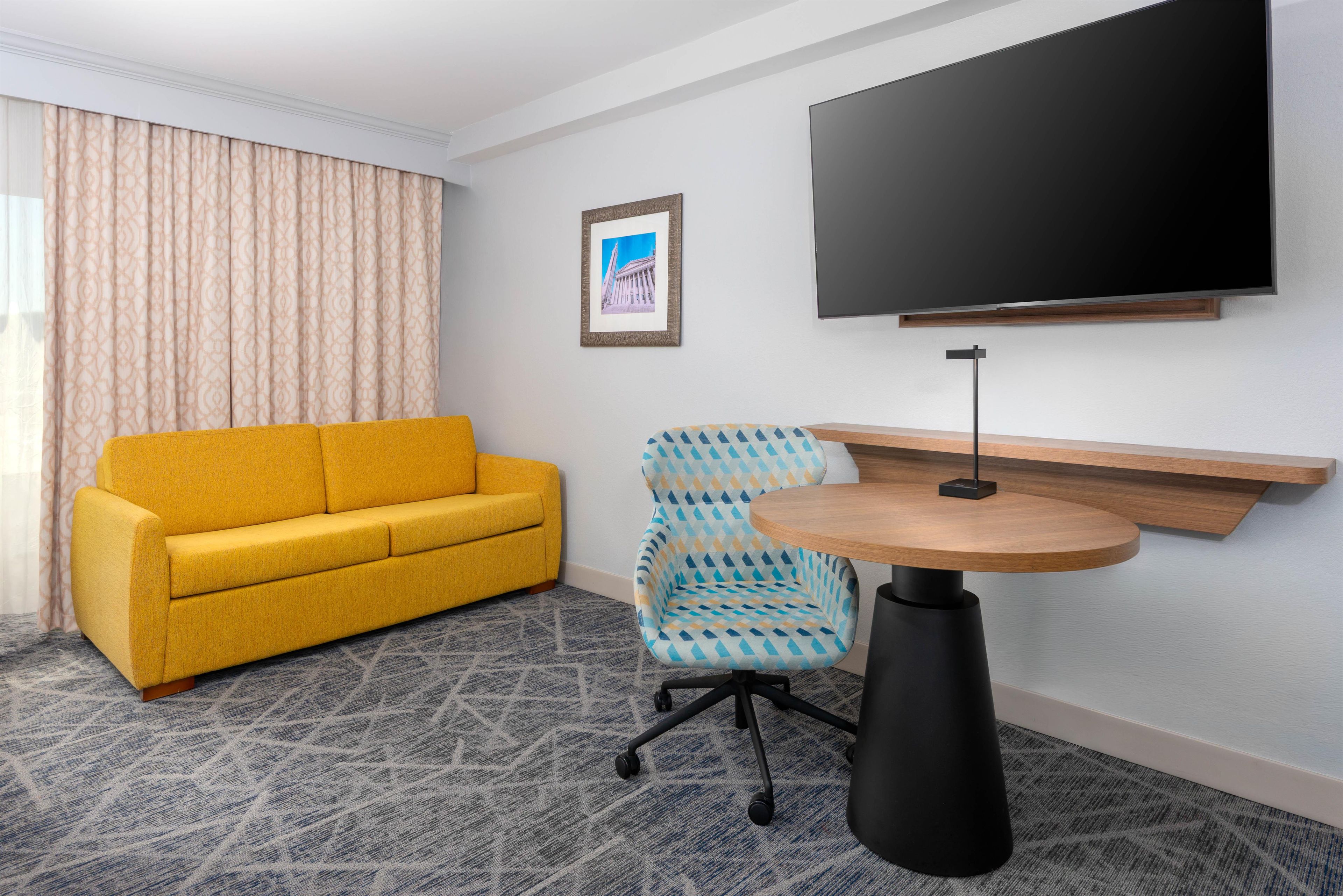 Habitación de hotel con sofá amarillo, un sillón estampado, una mesa redonda de madera, una lámpara de pie negra, un cuadro enmarcado en la pared y un televisor de pantalla plana grande montado sobre la mesa.