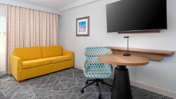 Habitación de hotel con sofá amarillo, un sillón estampado, una mesa redonda de madera, una lámpara de pie negra, un cuadro enmarcado en la pared y un televisor de pantalla plana grande montado sobre la mesa.