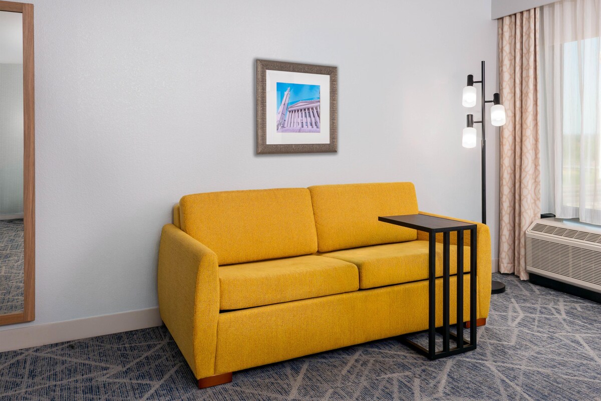 Habitación de hotel con un sofá amarillo, un cuadro enmarcado en la pared, una lámpara de pie y una ventana con cortinas.